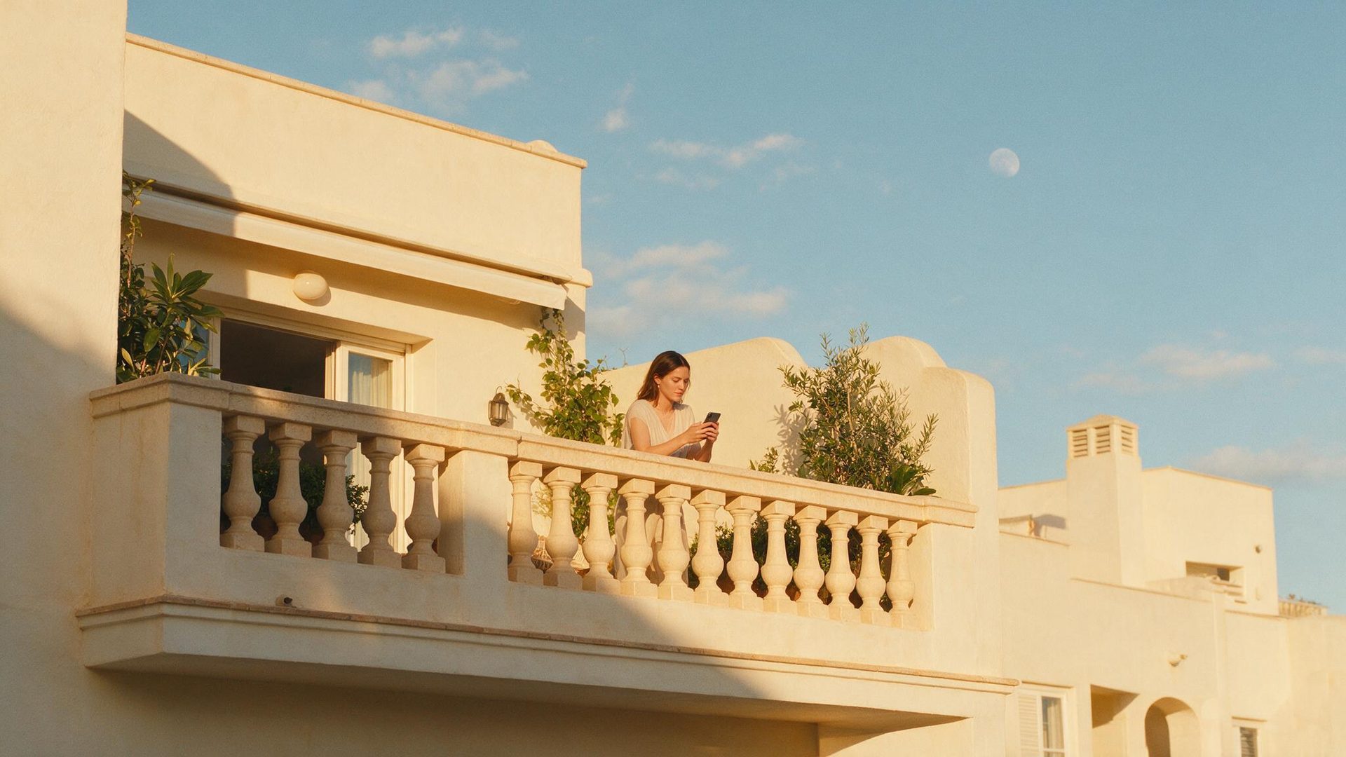 Mujer mirando su móvil en un balcón mediterráneo de piedra clara al atardecer, con una luna pálida visible en el cielo azul